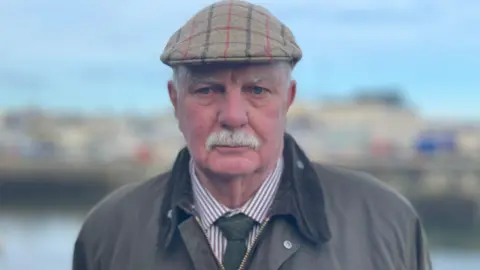 Stephen Carter looking ahead. He has a reflective expression on his face and is wearing a beige flat cap with a red and black tartan print, a khaki green coat with a dark collar, and a green tie and brown striped shirt.