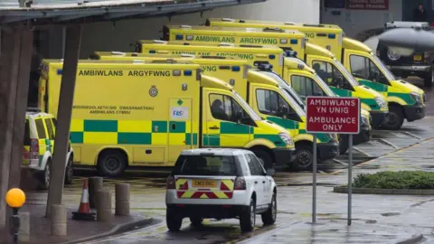 Five ambulances are lined up outside a hospital. Cars with emergency yellow and red zigzags are also visible. A sign says Ambiwans yn unig, Ambulance only. 