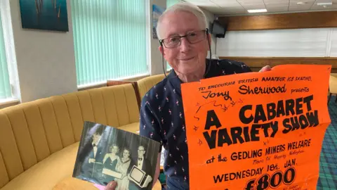 Tony Sherwood is looking into the camera and smiling. He is wearing glasses and holding an old poster which was used to promote a variety show at the then Gedling Miners Welfare Club. He is also holding a black and white photo of ice dancers Jayne Torvill and Christopher Dean. 