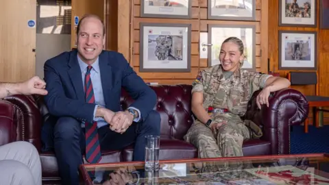 Welsh Guards Prince William sits on a burgundy, leather Chesterfield sofa in an army barracks, talking to someone sitting across from him. He is wearing a navy suit, light blue shirt and a navy and red tie. Sitting beside him is a girl in her early 20s who is wearing a camouflage army uniform with her blonde hair slicked back into a ponytail. She is smiling with her arm resting on the arm of the sofa. There are framed army photographs hanging on the wood panelled wall behind them.