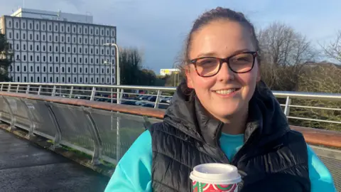 Cynthia is standing on a bridge and holding a disposable cup of coffee. She wears tortoiseshell glasses and has  a black body warmer on over a green jumper. Marlborough House is in the background.