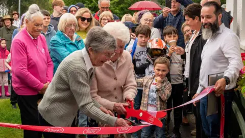 POST OFFICE Many people, young and old, gathered round to watch the ribbon-cutting ceremony for the opening of the Post Office. There is a red ribbon with the Post Office logo on it, and two ladies are using large red scissors to snip the ribbon.