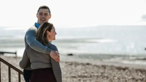 Peter Murphy has short black hair and is wearing a grey jumper. He has his arm around his wife, who has brown hair and is also wearing a grey jumper. They are standing by the beach in Shoeburyness.