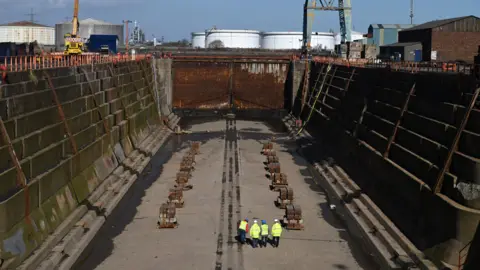 Four men standing inside a dry dock. They are wearing hard hats and hi-vis jackets. They appear very small because of the size of the dock's walls on either side of them. A large metal gate stands in front of them leading to the river.