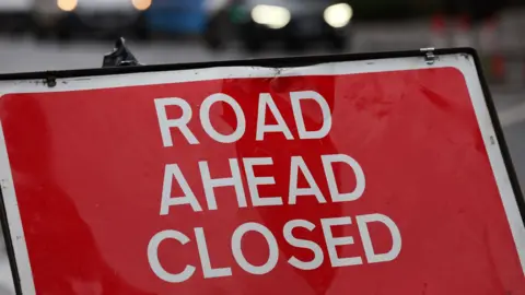 A red sign with white writing that reads ROAD AHEAD CLOSED.