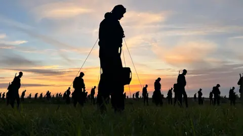 A large number of black silhouettes of servicemen in a garden. You can see grass in the front and a sunset behind. An airman figure is in the foreground. 