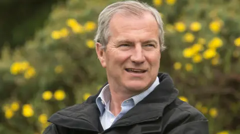Real Jardín Botánico de Edimburgo /PA Wire Foto de cabeza y hombros de Simon Milne, que tiene el pelo gris peinado en un cobertizo lateral. Viste una camisa a rayas azules y blancas y una chaqueta negra. Los arbustos y las flores amarillas son visibles sobre el fondo borroso. Mira a su izquierda y sonríe.