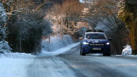 PA Media A car drives along a snow-covered road in Mold, Wales. There is snow on the hedges surround the road, which is partially-obscured by snow and ice.