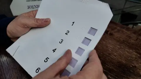 A close up of two hands holding the voting template. It seems to be a thin plastic sheet with the numbers one to six printed from top to bottom and a small open square beside each one where 