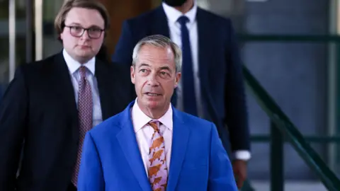 Nigel Farage walking outside, wearing a blue suit with a pink tie, decorated with a fish pattern. There are two men behind him, both wearing dark suits. 