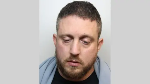 Custody image of Michael Elmer. he has a blue blanket around his neck and is looking down. He hs brown hair and a short, brown beard. 