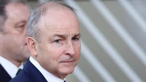 Reuters A close-up of Micheál Martin. He is an older man with thinning grey hair. He has a neutral expression on his face. He has a dark suit jacket and a white shirt.