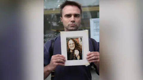 PA A worried looking man man with brown heair wearing a blue shirt is holding a photo of a woman with curly hair who is smiling at the camera.