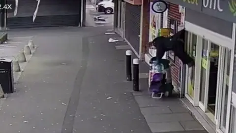 Lady hanging from the back of her coat outside a shop holding a trolley. She is horizontal and about four feet off the ground