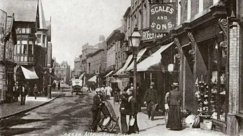A black-and-white period photograph of George Street in Altrincham from Trafford Council's archive. In the foreground a group of three men in suits can bee seen talking - one of them is holding a rocking chair. A woman in an ankle-length skirt, jacket and hat can be seen looking in a shop window. A horse and cart can be seen further up the street.