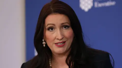 PA Northern Ireland Deputy First Minister Emma-Little Pengelly is speaking during a press conference. We can see the tops of her shoulders and head. She has shoulder-length dark hair and is wearing a pearl necklace and pearl earrings. She is wearing a dark jacket. In the background is a blue board with the logo of the Northern Ireland Executive