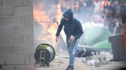 Getty Images A man holds a brick with a fire and scenes of disorder in the background.