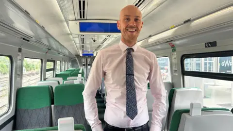 Adam Walding smiles at the camera while standing in a train carriage wearing shirt and tie