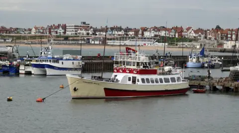 The Yorkshire Belle docked in Bridlington. It is cream and red with an enclosed seating area and upper deck. There are other boats and buildings in the background.