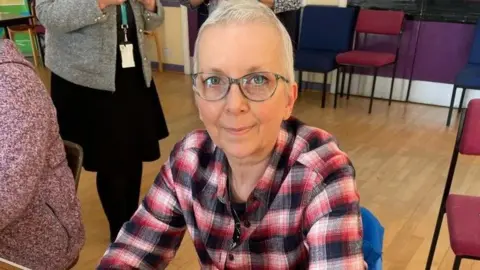 The Royal Wolverhampton NHS Trust Sharon Hever is wearing a red, white and black top. She has short white hair and is wearing glasses. Some chairs and people, whose faces cannot be seen, are in the background.