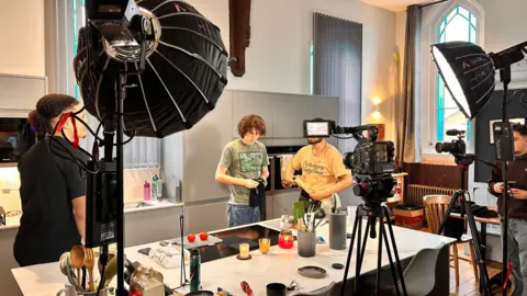 One in Two Behind-the-scenes view of a video shoot in a modern kitchen setup, with cameras, lighting equipment, and crew members filming two men at a kitchen island.