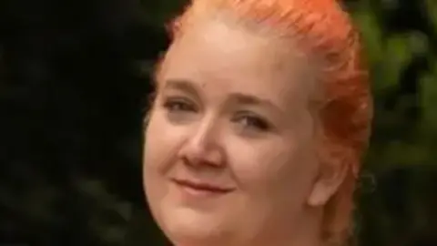 A close-up image of a woman, Rhiannon, who has red hair tied in a plait and is smiling at the camera.
