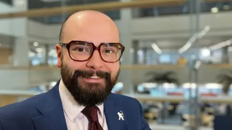 Man sits in an office in suit with a Robin Hood pin
