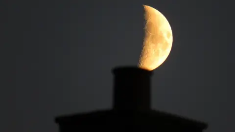 A quarter moon can be seen over the top of a chimney which is silhouetted