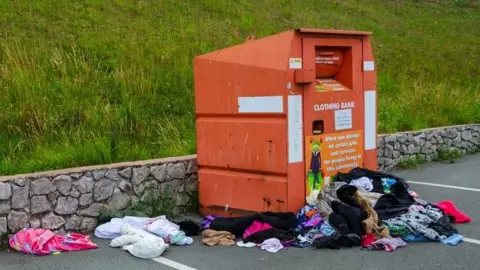 Getty Images A large orange recycling container with piles of clothes dumped in front of and around it.