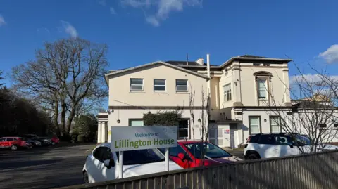 A large white building with several wings. There is a sign that reads "Lillington House" and a car park with vehicles in parking bays in front of the building. In the background is a large tree and the sky is blue.