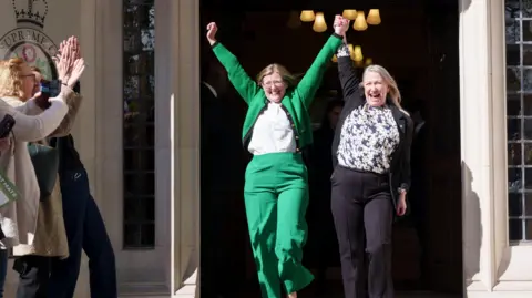 Getty Images Womens' rights campaigners with their arms raised in celebration outside a court. One woman has glasses and is wearing a green suit and white shirt. The other woman is earing a black jacket and white and blue floral shirt. They both have blonde or light brown hair. People are clapping at the side of the image.