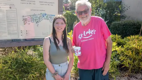 supplied An elderly bearded man in a pink t-shirt stands in the sunshine with a coffee mug. Next to him is a younger girl with dark hair wearing a green vest top.