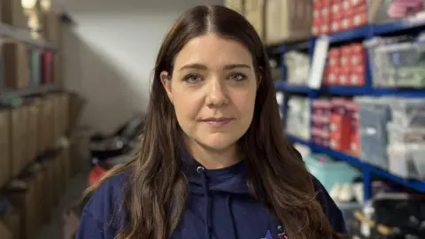 BBC A woman with long brown hair is wearing a dark blue hoodie. She is standing in front of shelves that are on the left and right of a narrow room. There are boxes and products stacked on them