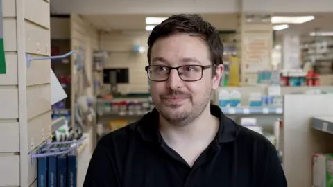 Shaun Whitmore/BBC Mark Jackson in a dark shirt and glasses standing in his pharmacy