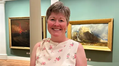 A woman with short brown hair stands in front of artwork smiling at the camera, she is wearing a cream sleeveless blouse with a rose pattern.
