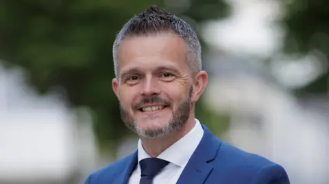 Robbie Butler. He has short grey-ish hair and beard, wearing a blue blazer, white shirt and navy tie.