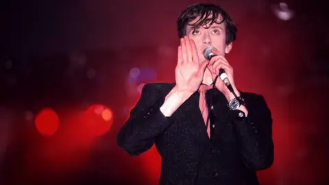 Jarvis Cocker pictured performing in 1995. He is wearing a dark suit, black shirt and pink tie.