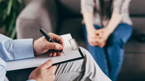 A man holding a clipboard perched on his knee. He is about to start writing, while in the background a woman sits on a sofa.