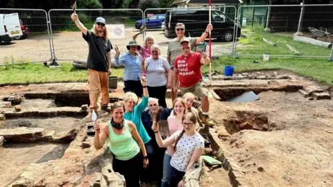 Oxford Cotswold Archaeology People between 17 and 78 smiling at the camera holding their spades while at the former mansion. 