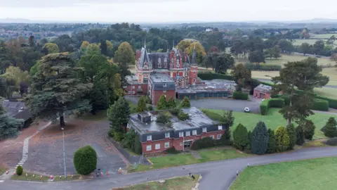 Greyfort Properties An aerial view of a hotel building. It is surrounded by trees and open countryside. The main building has a number of turrets and conical towers. There is a separate building, which is smaller and has a flat roof.