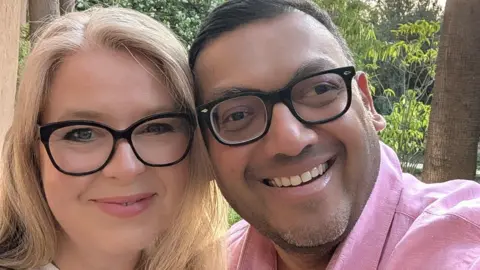 HANDOUT A smiling blonde woman wearing glasses is seen cuddled up next to a smiling man with short brown hair also wearing glasses in a selfie 