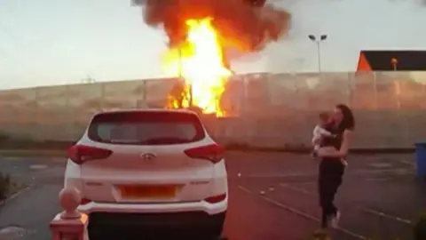 BBC A fire takes hold in a fence, as a woman carrying a baby runs to a white car parked in a car park, opposite the fire