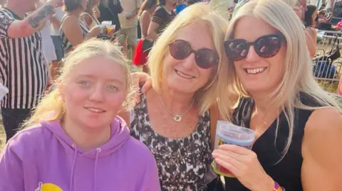 Helen smiling before she became unwell with her daughter and two granddaughters, wearing sunglasses and a leopard print top. They are stood at a festival in the sun, with people in the background. 