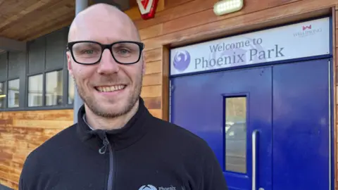 A man with a bald head and stubble beard smiling at the camera. He has black rimmed glasses and is standing in front of a blue door that has the sign 'Welcome to Phoenix Park' above it.