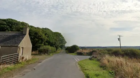A rural road with a farm building on the left side of the image. There is a junction, and further up the road a small wooded area.