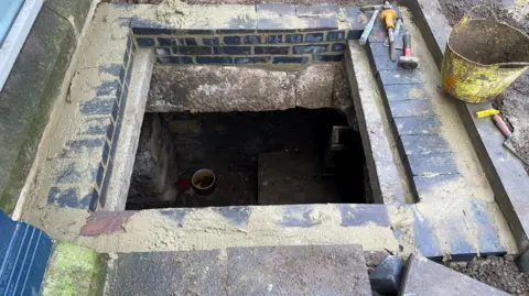 A square brick-lined opening to a cellar is shown with tools on one side and a yellow bucket 