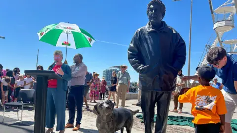 The statue in full showing Morgan with his hands in the pockets of his coat and a Labrador dog beside him. There is a young boy looking up at it, with many others beside the statue on the right. On the left a woman is giving a speech at a podium with a man holding a Wales flag umbrella over her head on a blue sky sunny day. 