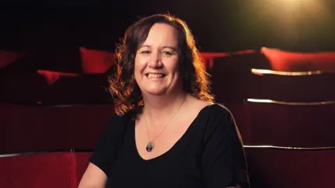 The Seagull Karen Read sits in a row of theatre seats and smiles at the camera. She has long, curly dark red hair and wears a black T-shir with a necklace resting on her chest. 
