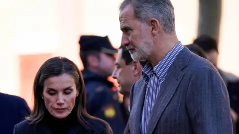 Reuters Queen Letizia and Spain's King Felipe arrive to Reina Sofia hospital in Cordoba. Letiza (on the left) has dark hair and is dressed in a long dark coat. The king has a grey suit on and an open-necked stripy shirt.