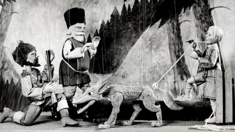 National Puppetry Archive A black and white photograph of puppets on a stage, suspended by string. There are two men, one with a long-barrelled gun, a boy and a wolf.